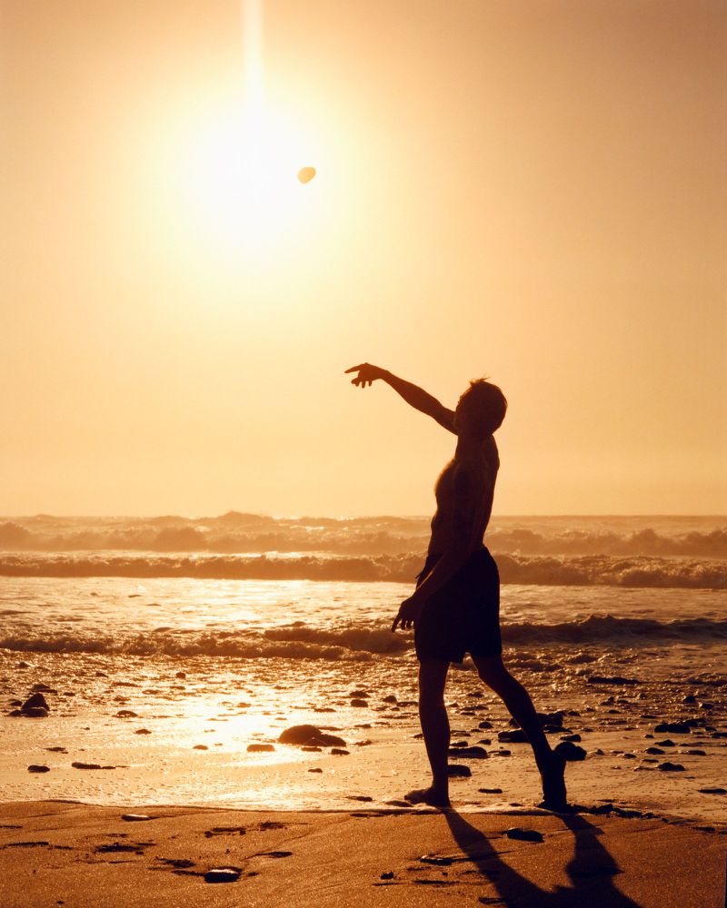 Silhouette of a man throwing a pebble into the air on a sandy beach during golden hour sunset by the ocean.