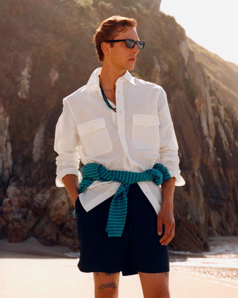 Man wearing sunglasses, a white shirt, and dark shorts outdoors on a sandy beach with rocky cliffs