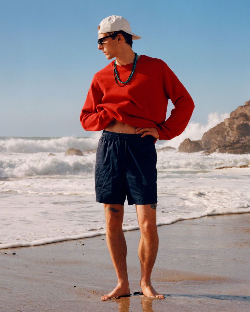 Model Quentin Demeester poses barefoot on a sandy beach wearing a bright red sweatshirt, navy swim shorts, and a white cap.