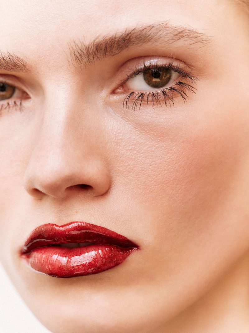 Close-up beauty portrait of a model with glossy red lips and defined dark mascara eyelashes against a neutral background.