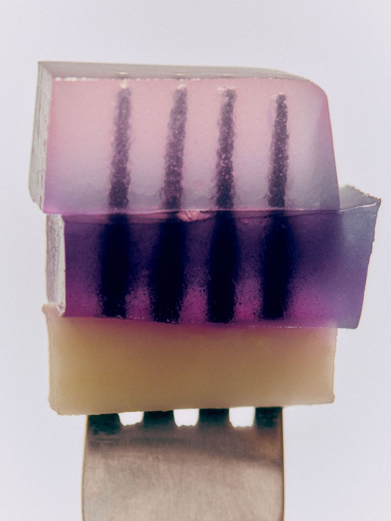 A layered translucent pink and purple gelatin dessert with vertical dark patterns, served on a metal fork against a white backdrop.