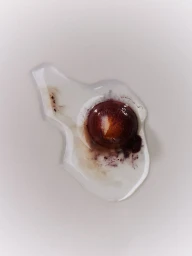 Dark red cosmetic sphere surrounded by a pool of clear liquid with bleeding pigment on a clean light surface.
