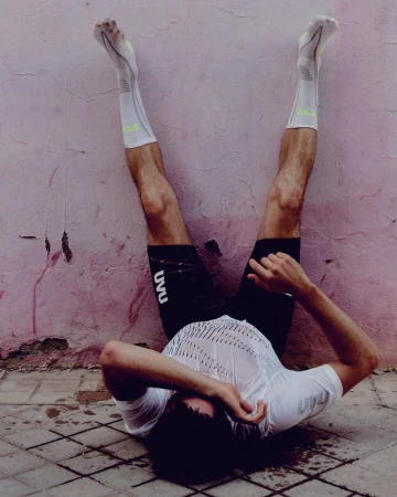 Athlete wearing white UVU athletic shirt and black shorts lying on tiled ground with legs propped up against a pink textured wall.