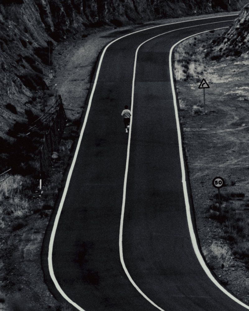 Monochrome high-angle shot of a lone runner moving down a winding asphalt road flanked by rugged hills and traffic signs.