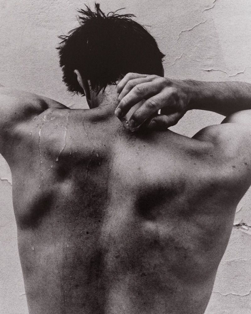 Monochrome back view of a man with water droplets running down his bare skin, holding his neck against a textured wall.