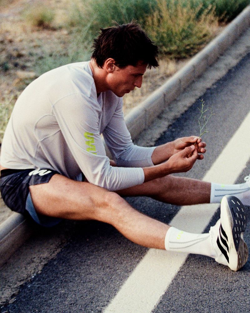 A male runner in a long-sleeve athletic top and running shorts sits resting on an asphalt roadside curb.