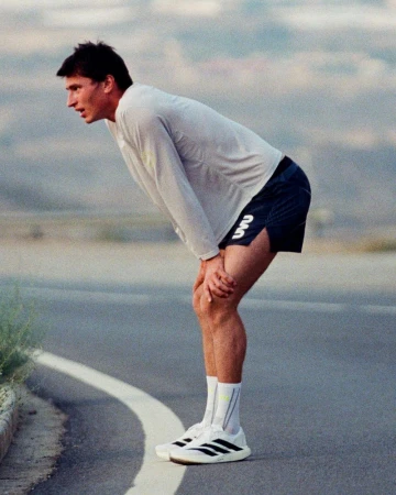 Male athlete in a light long-sleeve running shirt and dark shorts resting with hands on his knees along a curved road.
