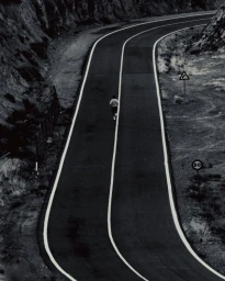 Monochrome high-angle shot of a lone runner moving down a winding asphalt road flanked by rugged hills and traffic signs.