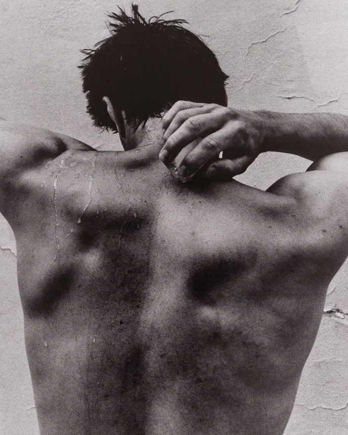 Monochrome back view of a man with water droplets running down his bare skin, holding his neck against a textured wall.