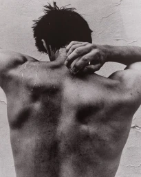 Monochrome back view of a man with water droplets running down his bare skin, holding his neck against a textured wall.
