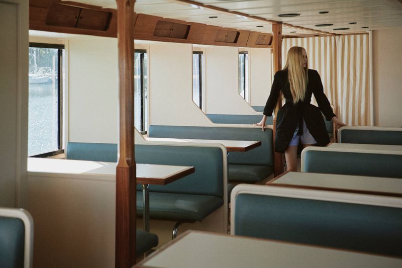 Model Maelle Swaelens standing in a retro passenger ferry cabin wearing a black belted coat.
