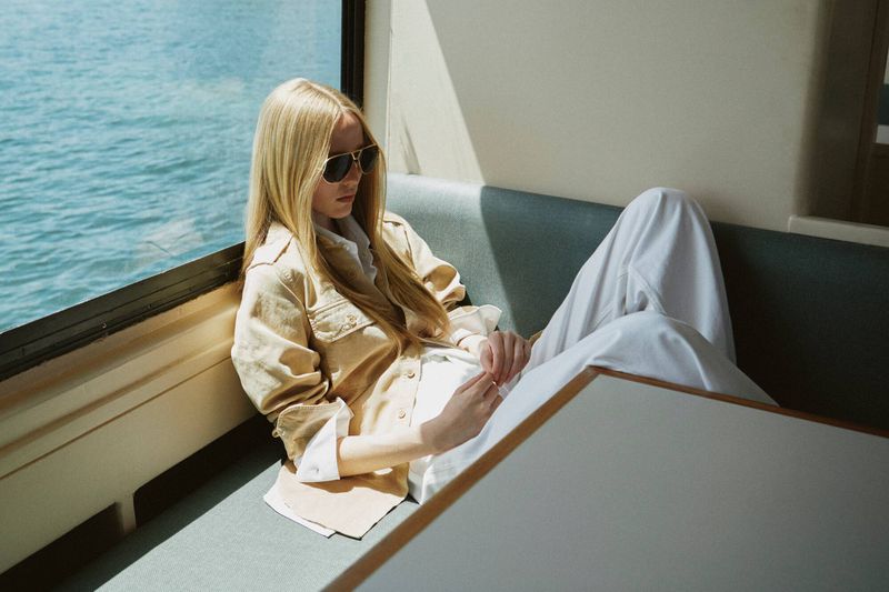 Model Maelle Swaelens wearing aviator sunglasses, a beige jacket, and white trousers while lounging by a window over open water.