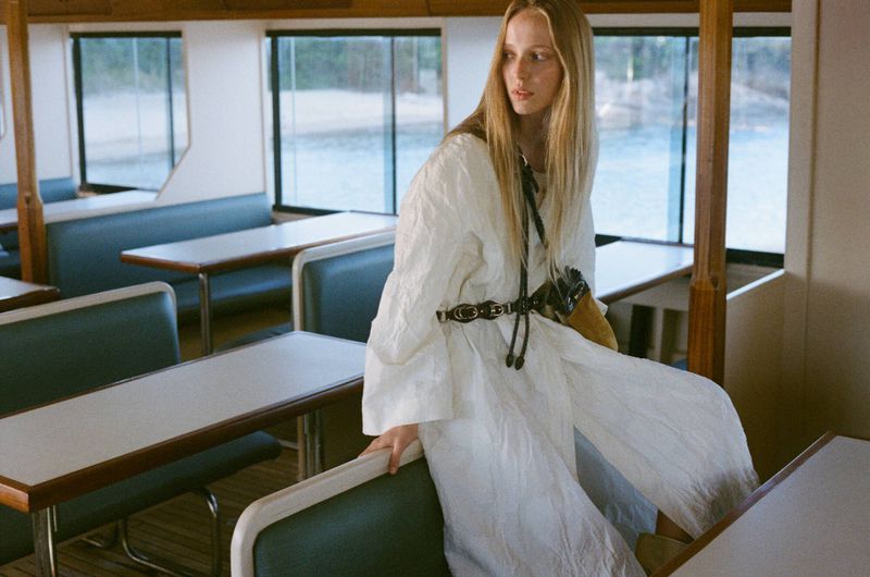 Model Maelle Swaelens wearing a textured white belted dress while seated in a passenger boat cabin with teal booth seating.