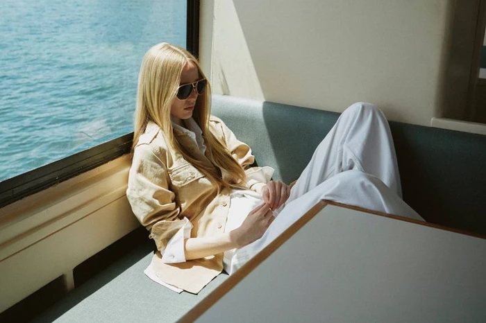 Model Maelle Swaelens wearing aviator sunglasses, a beige jacket, and white trousers while lounging by a window over open water.