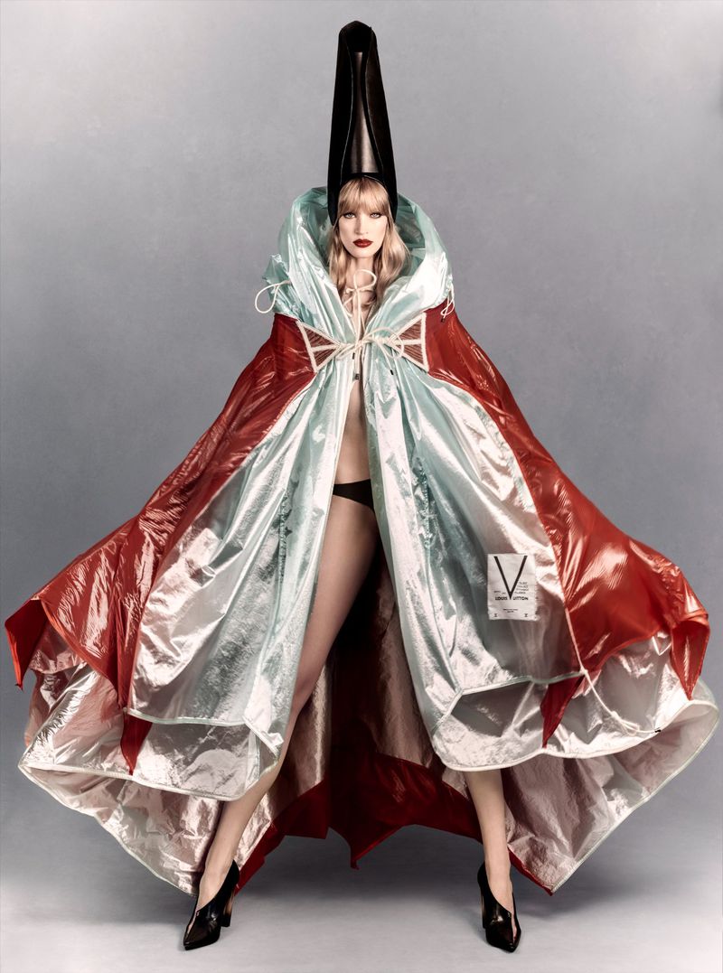 Model Jacqui Hooper in an oversized red and silver metallic hooded cape and tall black conical headpiece against a grey studio backdrop.