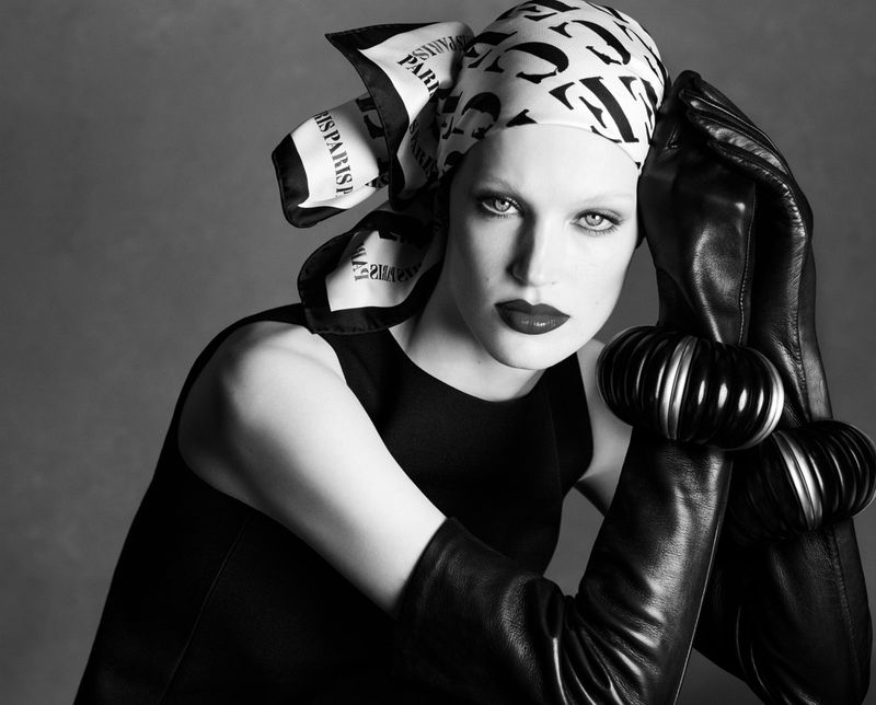 Model Jacqui Hooper wearing a black and white patterned headscarf, long black leather gloves, and chunky bangles against a grey backdrop.