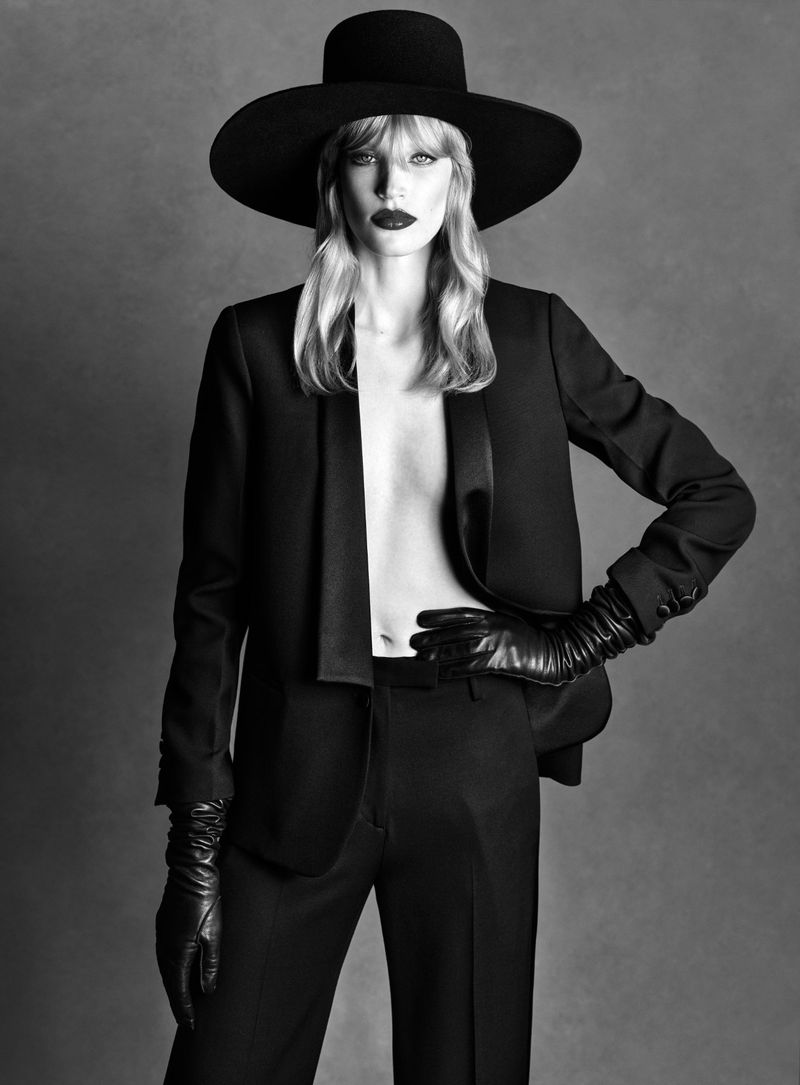 Model Jacqui Hooper posing in an open black blazer, matching trousers, wide-brim hat, and leather gloves against a studio backdrop.
