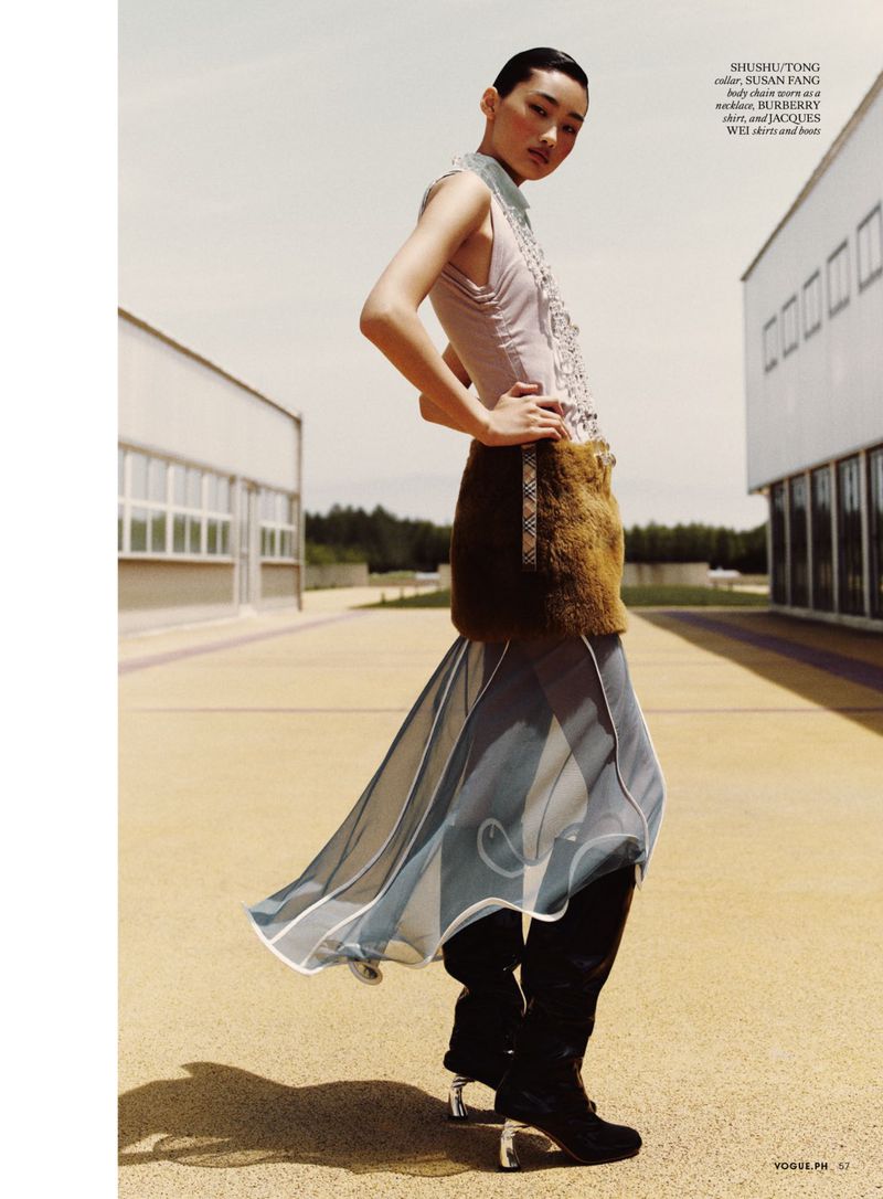 Fashion editorial shot featuring a woman in avant-garde sleeveless top, skirt, boots in an outdoor setting with modern building.