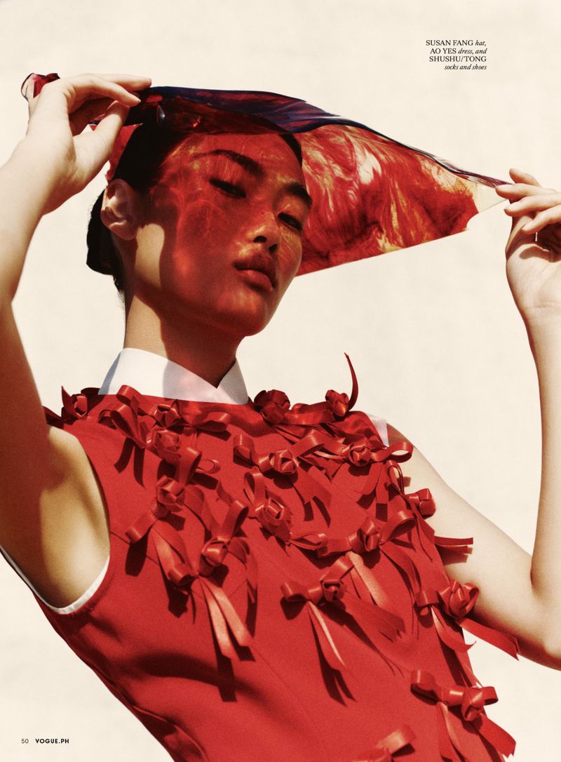 Fashion editorial shot of a woman wearing a red dress adorned with bows and a sheer red head covering against a light background.