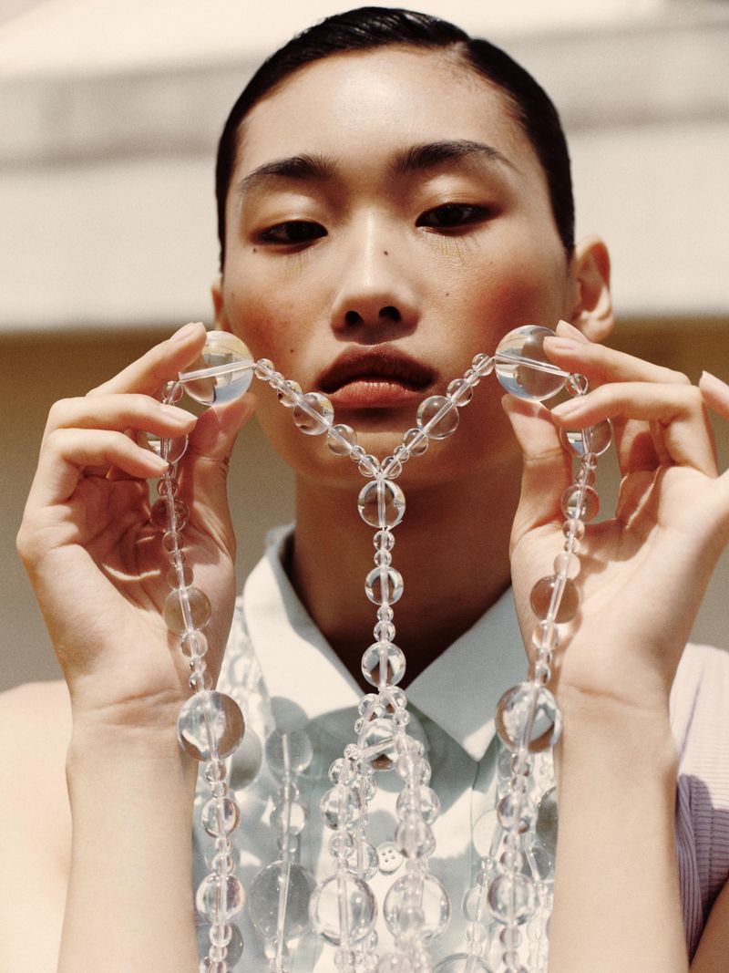 Asian woman poses with clear beaded necklace against a neutral background, wearing a collared shirt and natural makeup look.