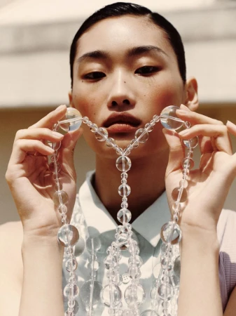 Asian woman poses with clear beaded necklace against a neutral background, wearing a collared shirt and natural makeup look.