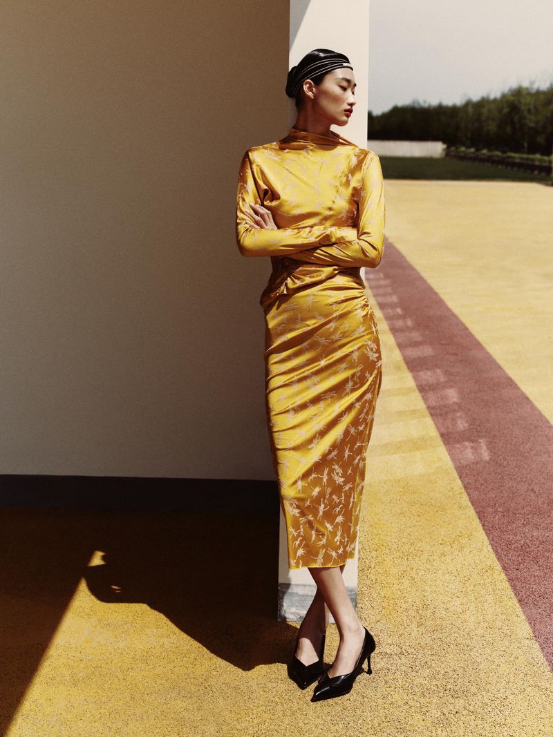 Elegant Asian woman in a gold dress poses outdoors against a textured background with geometric patterns in warm afternoon light.