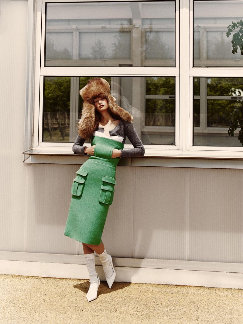 A woman in a fur hat, green dress, and white boots leans against a windowed wall outdoors, capturing a blend of high fashion and urban style.