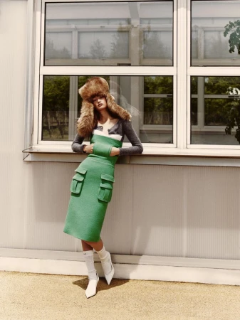 A woman in a fur hat, green dress, and white boots leans against a windowed wall outdoors, capturing a blend of high fashion and urban style.