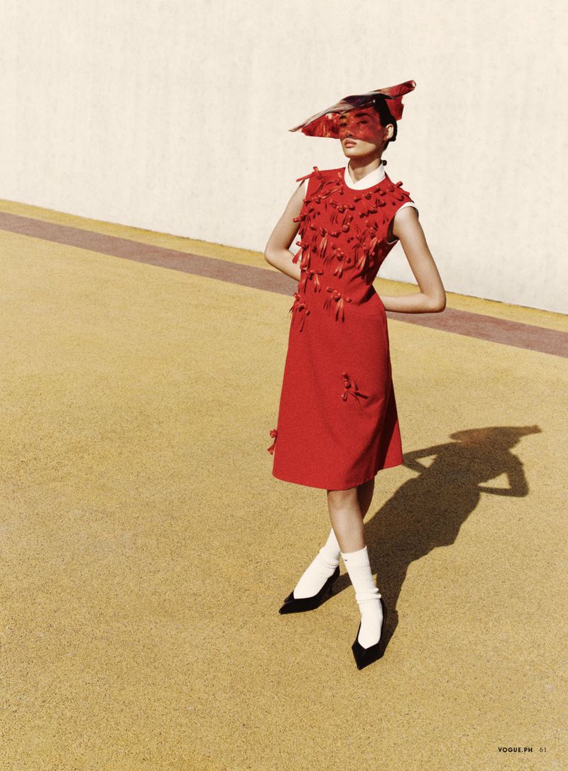 A fashionable woman in a red dress and a red headpiece posing outdoors on a yellow surface with a white wall in the background.