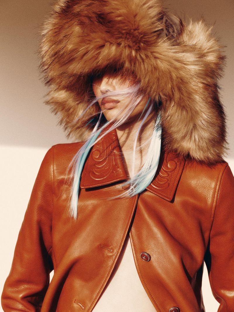 Fashionable adult woman with blue-streaked hair wears a fur hat and leather jacket in a close-up portrait against a neutral backdrop.
