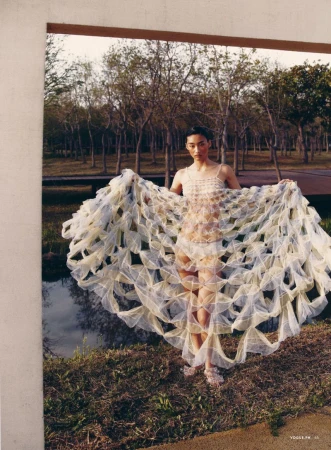 Fashion shot of an Asian female model in a park, wearing a cream-colored, geometrically patterned gown, standing near a pond surrounded by trees.