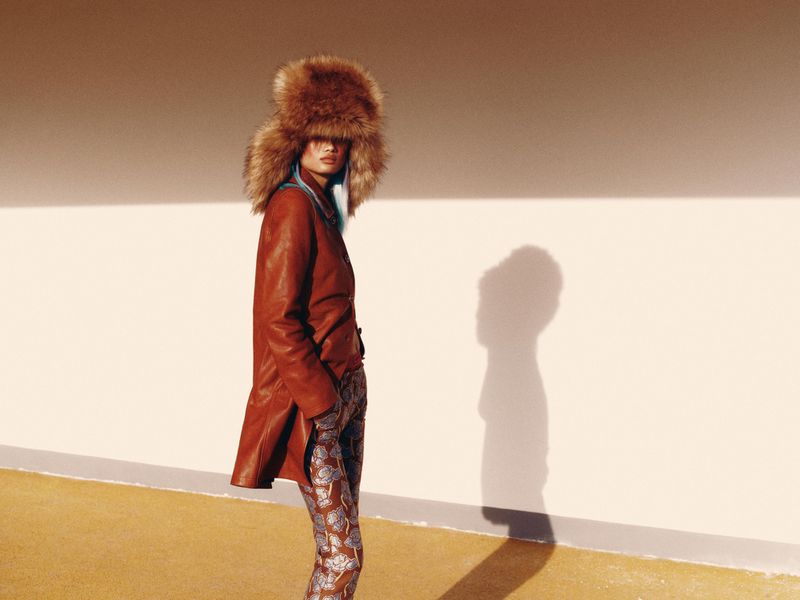 A woman poses wearing a brown fur hat, jacket, and patterned pants against a white wall in a fashion editorial shot with natural light.