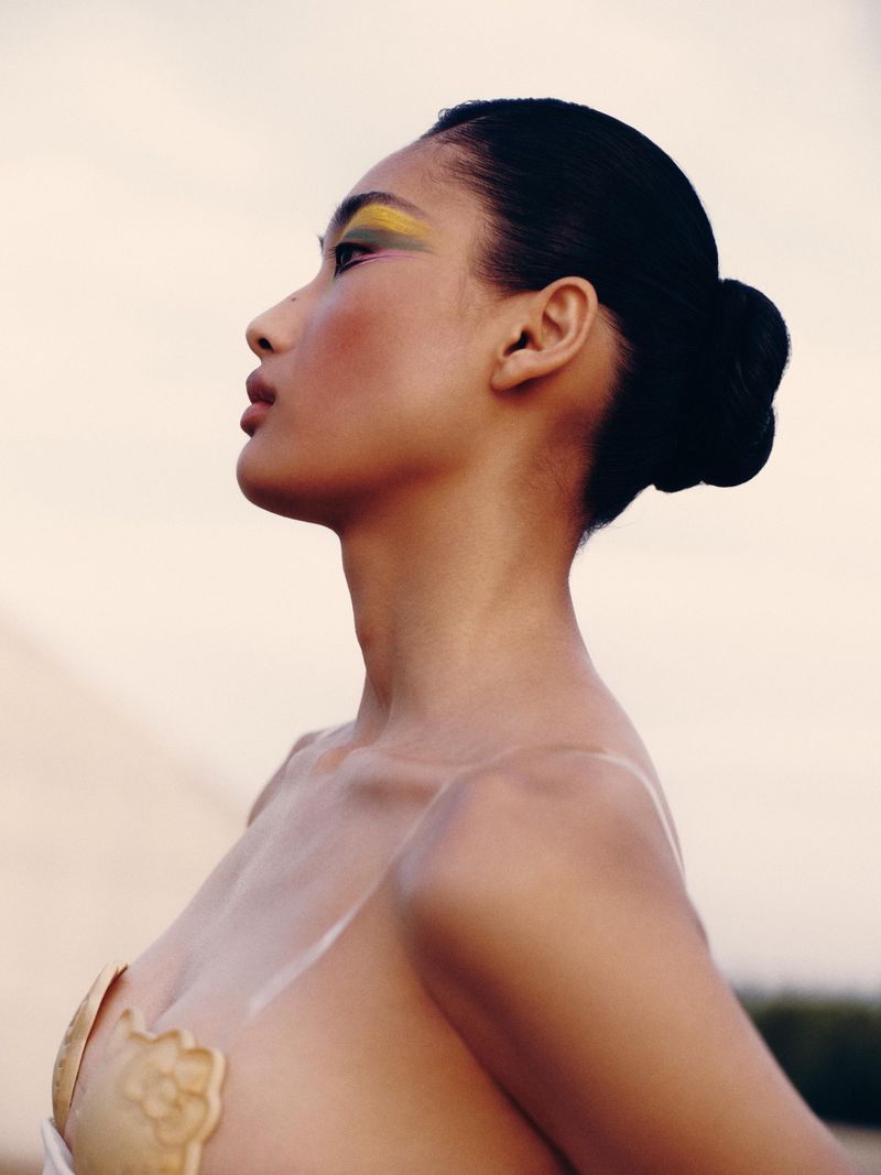 Profile shot of an adult woman with bright yellow and green eyeshadow, wearing clear shoulder straps and a textured tan garment.