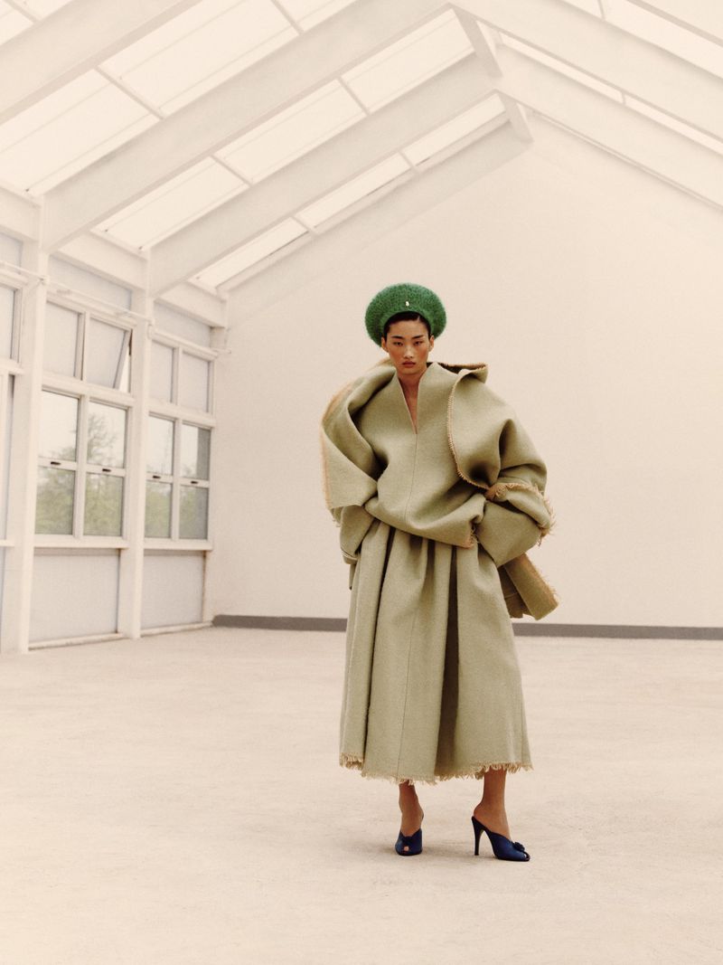 A stylish Asian female poses indoors wearing an oversized, earth-toned outfit with a green hat and blue heels in a minimalist setting.