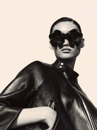 Model Xiuli Jiang wearing oversized black floral sunglasses and a structured leather jacket against a warm neutral background.