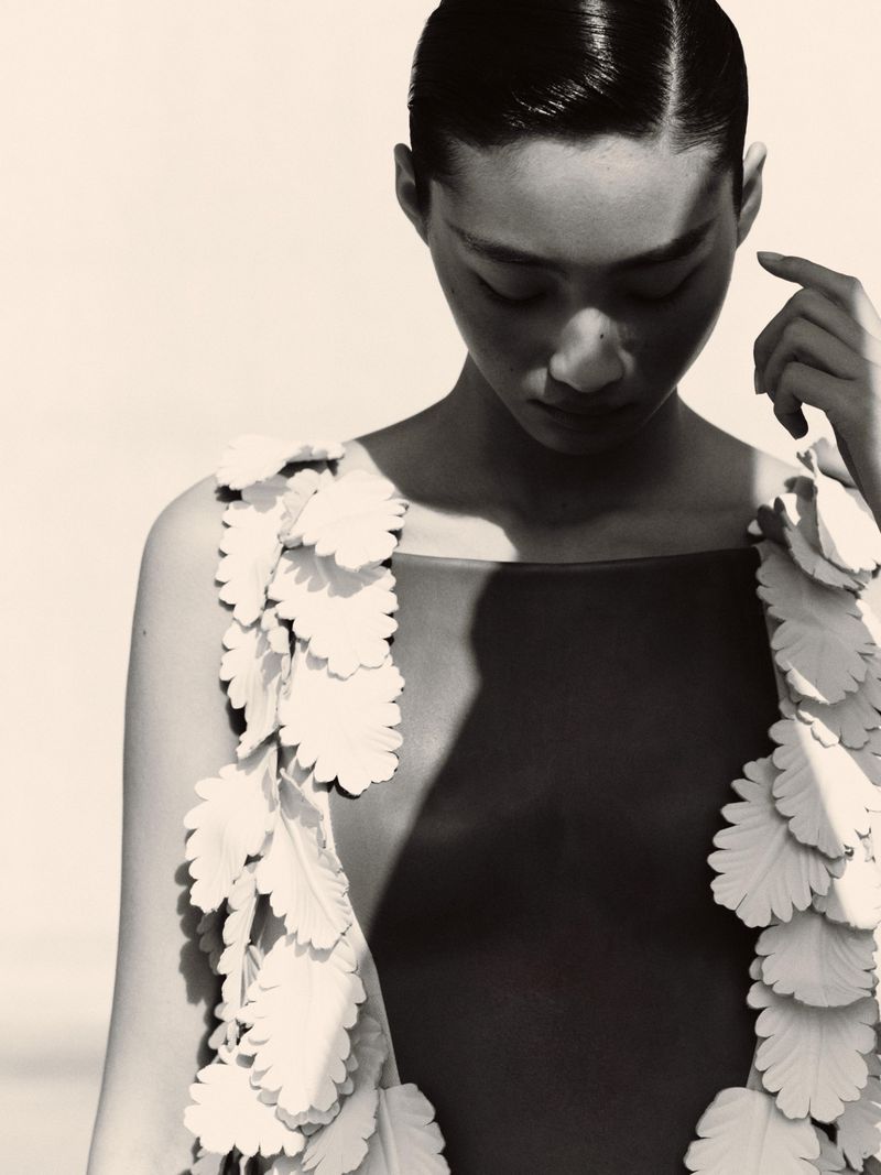 Monochrome portrait of an Asian woman with sleek hair, wearing a dark top adorned with layered white floral accents on the shoulders.