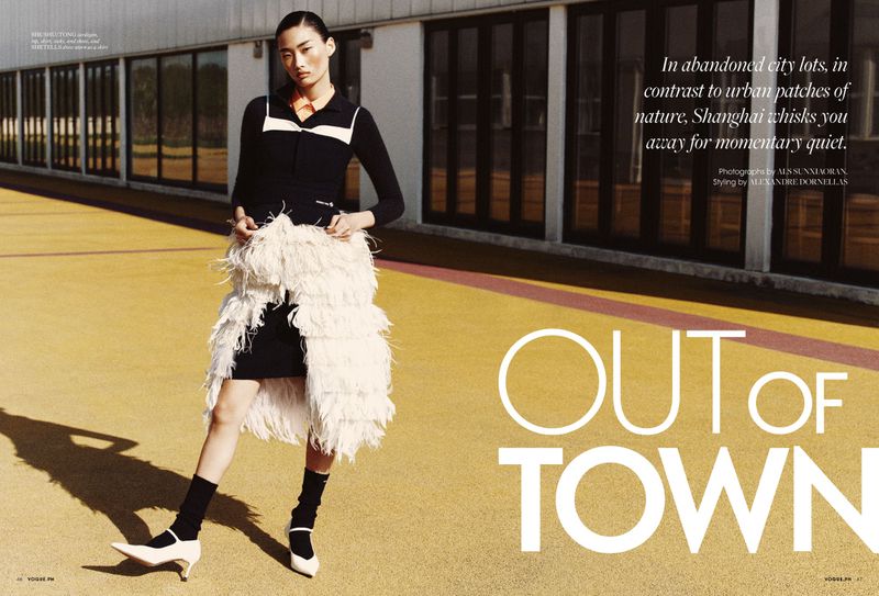Fashion editorial shot of an Asian female model in a black dress with a layered white fringe coat, posing on a yellow outdoor surface.