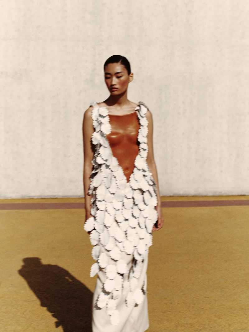 Asian woman in an elaborate white dress with petal-like details and a deep-V cut, posing in front of a yellow wall.