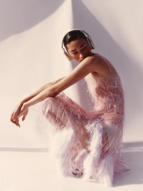 Asian woman in a fringed pink dress and headphones poses in a sunlit studio, casting soft shadows on the white backdrop and floor.
