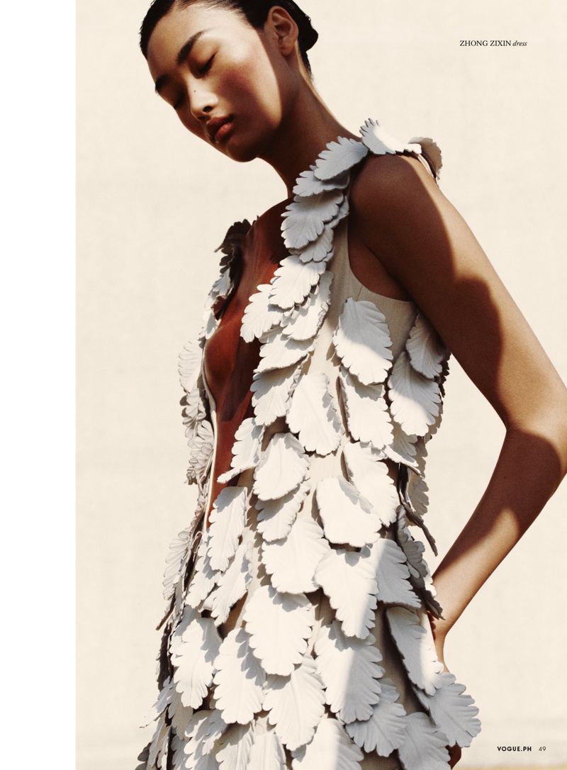 Fashion shot of an Asian adult female model wearing a sleeveless dress adorned with white leaf-like decorations, set against a light background.