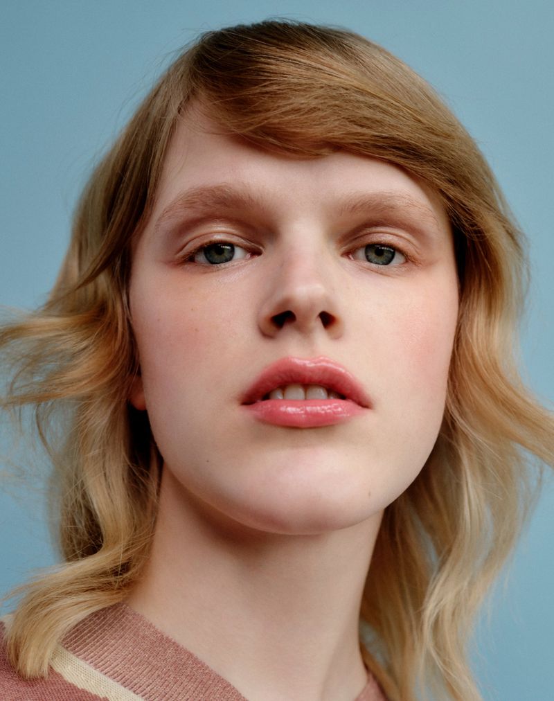 Close-up portrait of a model with blonde hair and glossy pink lips against a plain blue background.