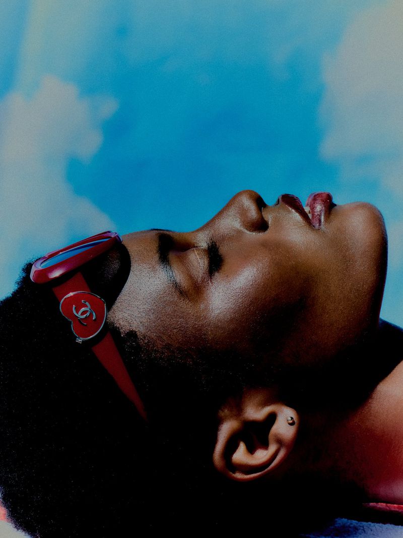 Close-up profile of a Black woman wearing red heart-accented sunglasses pushed back on her head against a blue sky.