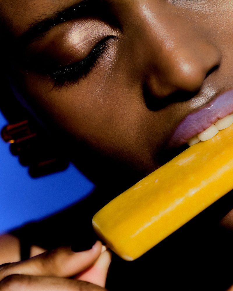 Extreme close-up of a woman with dewy skin and glossy purple lips biting a yellow popsicle against a solid blue background.