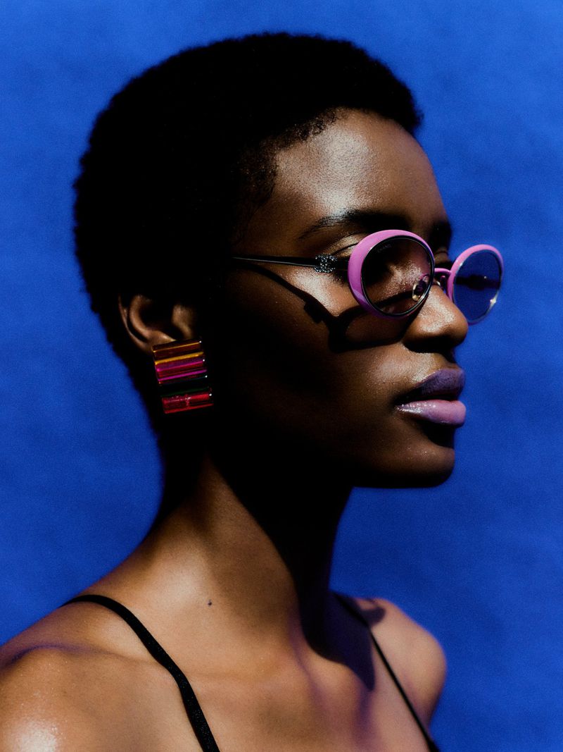 Close-up portrait of a woman wearing pink oval sunglasses and multi-colored block earrings against a vivid blue background.