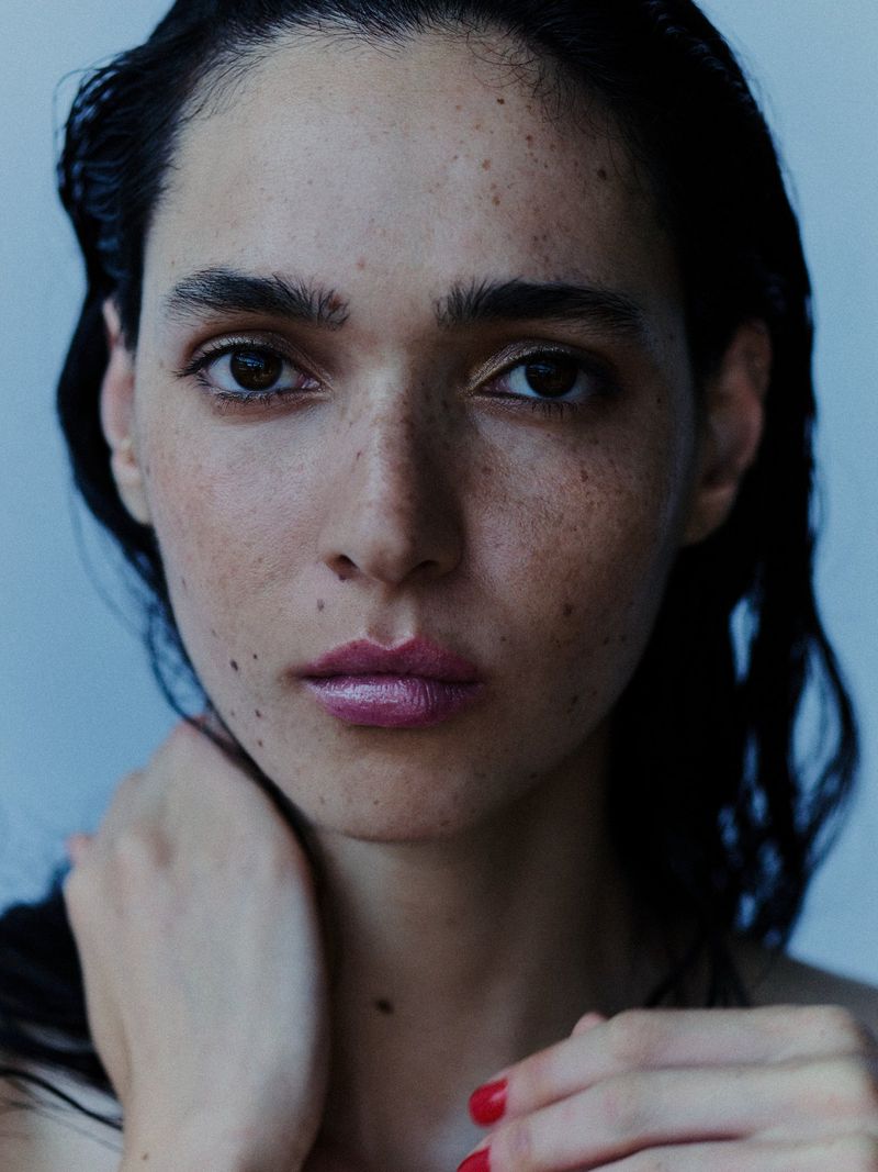 Close-up beauty portrait of a woman with freckles, wet dark hair, pink lipstick, and red nail polish looking directly at the camera.