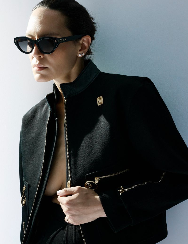 Model Laura Rylands wearing black sunglasses and a black structured zip-up jacket against a light backdrop.