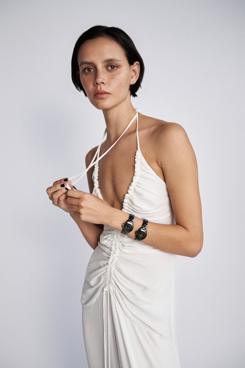Woman in a ruched white halter dress wearing two black wristwatches on one wrist against a light seamless studio backdrop.