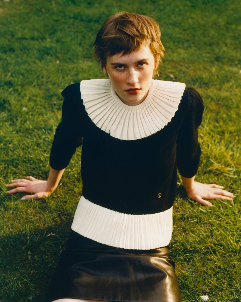 Woman with short brown hair seated on grass wearing a black knit sweater with a white pleated collar and black leather skirt.