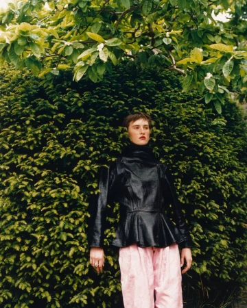 Model wearing a black leather peplum jacket and pastel pink trousers standing before a dense green hedge with leafy branches above.