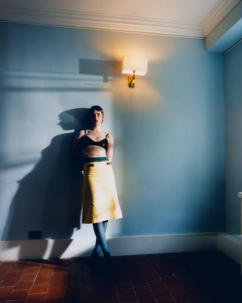 Model wearing a black bralette and yellow midi skirt leaning against a pale blue wall under a sconce light.